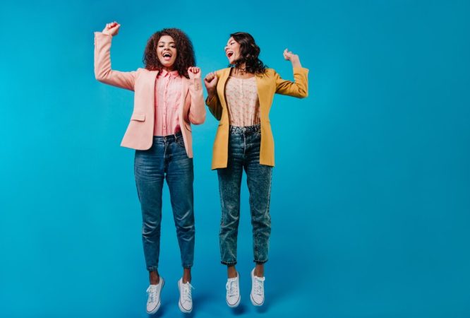 two-women-jeans-jumping-blue-wall_197531-12950
